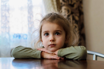 a little girl with big brown eyes and long eyelashes dressed in a T-shirt of khaki color put her hands on a brown wooden table, put her head on her hands and looks forward