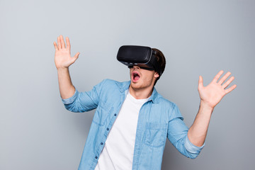 Close up of impressed, shocked, handsome, attractive guy in jeans shirt wearing virtual reality goggles, getting experience using VR-headset, gesticulating, gesturing with hands  over gray background