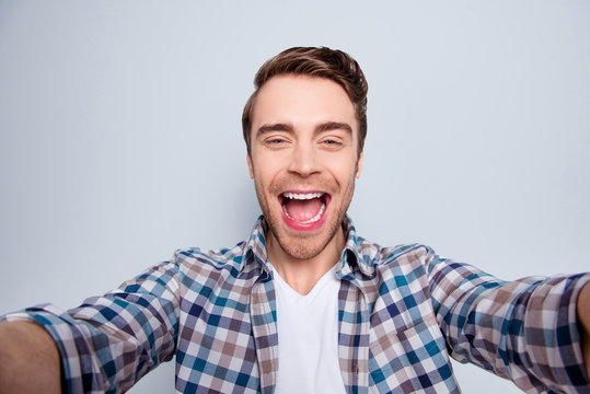 Self portrait of bearded, cheerful, funny, happy guy in casual outfit, checkered shirt with wide open mouth, laughing over grey background