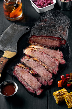 Fresh Brisket BBQ beef sliced for serving against a dark background with sauce, hot peppers and corn.