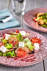 Healthy mixed Greek salad served on a pink plate with silver fork containing crisp leafy greens, microgreen, feta, onion, tomato and sliced beef