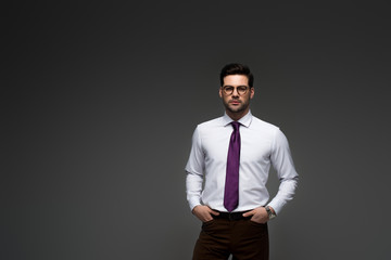 Businessman in glasses standing with fingers in pockets isolated on grey