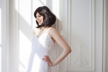 Portrait of young attractive brunette with short hair in a wedding dress. Standing at the white walls. Joyful bride.