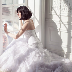 Portrait of young attractive brunette with short hair in a wedding dress. Sitting near the window, back view.