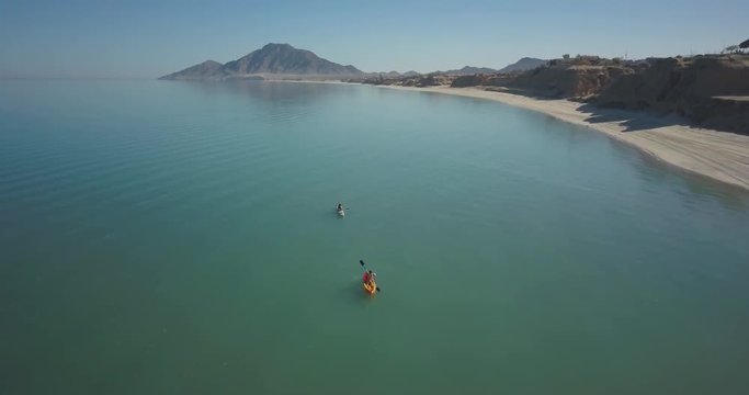 Aerial camera high flying backwards as two female kayakers paddle forward in a calm sea.