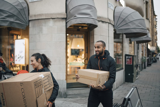 Smiling male and female workers carrying cardboard boxes while walking in city during delivery