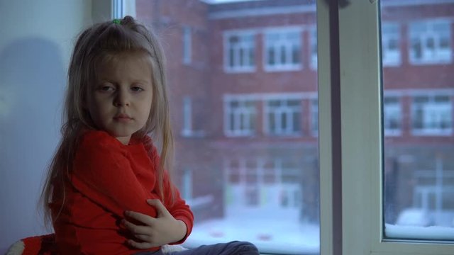 A cute little girl in a red T-shirt with a long sleeve is sitting on the windowsill looking at the snowy landscape.