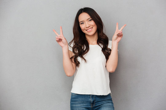 Portrait of asian attractive woman in casual t-shirt and jeans smiling and showing victory sign with both hands, isolated over gray background