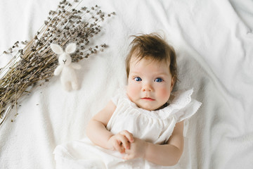 Gorgeous child in white dress lies on white bed with tiny rabbit in a bright room