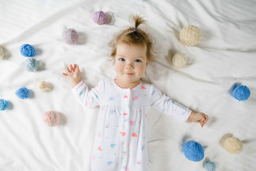 Pretty little girl lies among colorful knitting balls on the white bed
