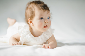 Gorgeous child with blue eyes in white dress lies on white bed in a bright room