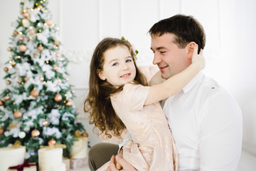 Obraz na płótnie Canvas Happy father holds his little pretty girl in peach dress standing before a Christmas tree