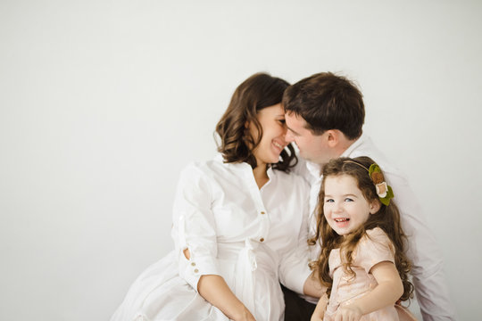 Pregnant woman, her man and their cheerful little daughter in peach dress hug each other tender sitting before a white wall