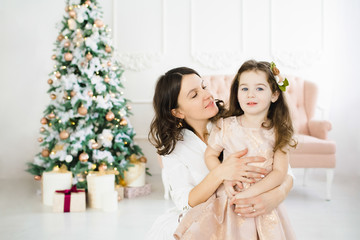 Obraz na płótnie Canvas Charming mom and her pretty little daughter pose before a rich Christmas tree in a luxury white room