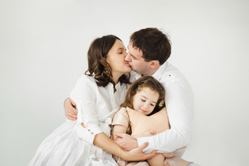 Fototapeta na wymiar Pregnant woman, her man and their cheerful little daughter in peach dress hug each other tender sitting before a white wall