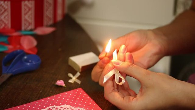 A woman cauterizes with a match of the edge of a satin ribbon, from which a bow for a postcard is made.