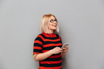 Fototapeta premium Smiling middle-aged blonde woman in sweater and eyeglasses holding smartphone