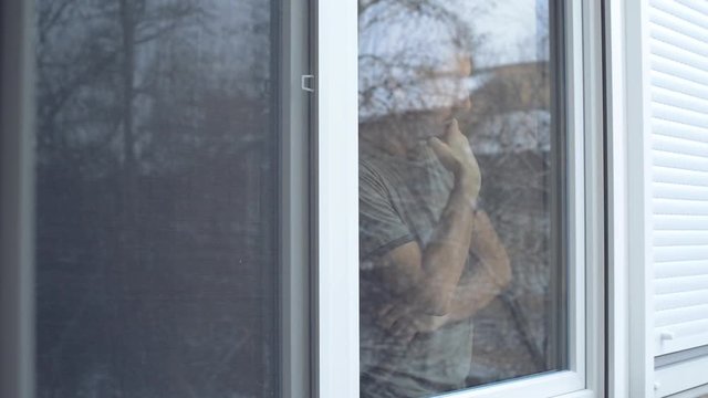 Depressed Man Standing By The Window And Looking Outside At Snow Falling In Cold Winter Afternoon, Teal And Orange Toned Footage With Selective Focus