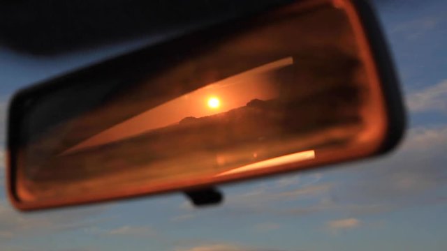 Retrovisor interior de coche en movimiento reflejando una puesta de sol anaranjada en contraste con la vista frontal de cielo azul con nubes. Cruzando un puente o paso elevado