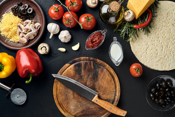 Cutting wooden board with traditional pizza preparation ingredients: cheese, tomatoes sauce, basil, olive oil, pepper, spices. Black texture table background
