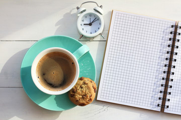 Coffee in a turquoise mug and cookies near a notebook on a white table and the white alarm clock...