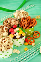 Popcorn, multicoloured drops, pretzels with salt and pistachio nuts in paper cups on green background. Snacks assortment