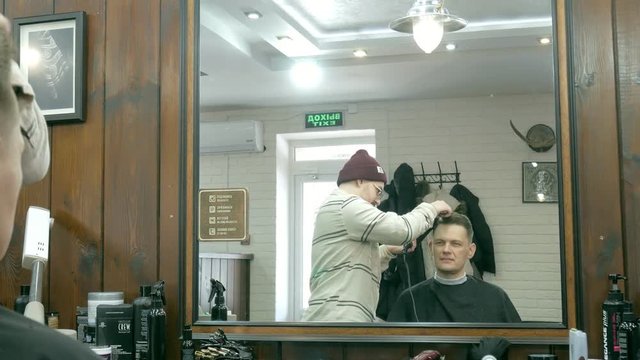 Barber getting groomed at man with hair dryer while sitting in chair at barbershop