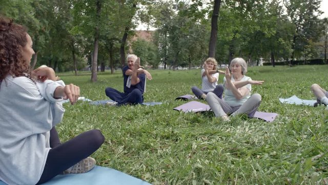 Group of six retired people sitting on grass on yoga mats and doing arm and shoulder stretches with their young female trainer