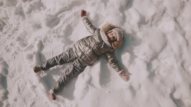 A little girl makes snow angels in snow. Top view.