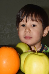 The little boy brown-eyed brunette sits at a table with apples and oranges, is going to eat and dreams