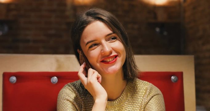 Brunette pretty young women portrait in gold sweater and red lips alks by phone. Loft style cafe background. Evening lighting.