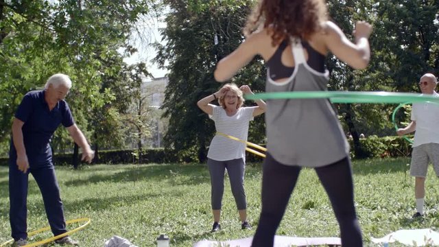 Seniors and their young female trainer spinning hula hoops during workout in park on warm summer day