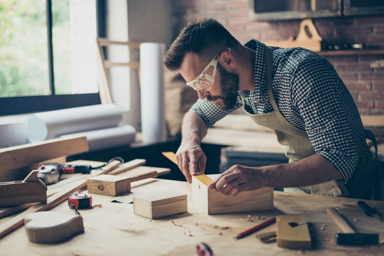 Concentrated persistent talented professional carpenter bearded wearing casual checkered shirt apron safety glasses is measuring the length on wooden box's side using a ruler