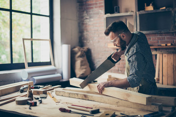 Concentrated strong confident professional handsome bearded wearing checkered shirt apron protective glasses is sawing a wooden plank with a handsaw in his workshop
