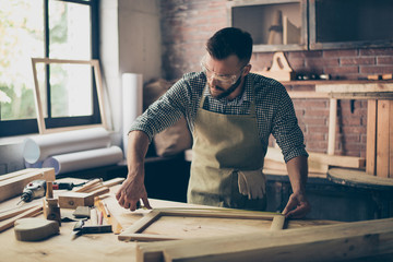 Busy skilled masterful experienced confident bearded dressed in checkered shirt and apron is taking wooden frame's measurements