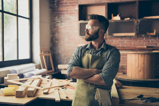Portrait of pensive handsome bearded minded calm confident experienced carpenter standing in front of his work-table with folded arms, he is looking aside