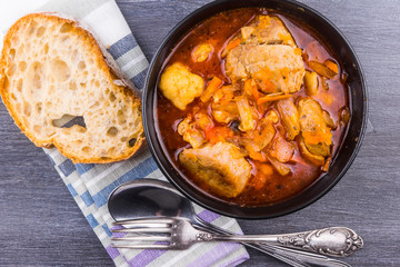 Stewed pork with vegetables in a black bowl and a piece of bread on a rustic table close up - top view