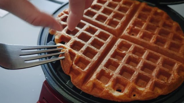 Girl removes golden brown Belgian waffles from a waffle iron in 4k. The teen uses a fork to pry the sweet baked pastry from the hot plates of the waffle maker. The breakfast food is a family favorite.