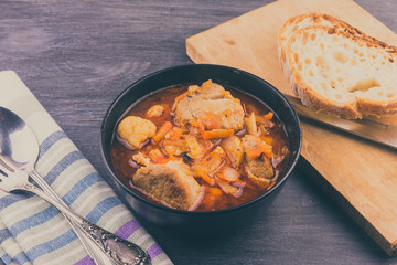 Braised pork with vegetables in a black bowl and a piece of bread on a rustic table