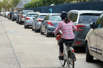 A woman in city clothes in a hat rides a bicycle along parked cars on a Paris street