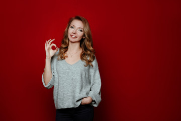 Body language and emotions.Portrait of beautiful cheerful blonde girl with curly hair over red background.The woman makes ok gesture and smiles at camera, has happy expression