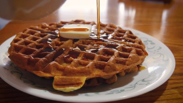 Pouring maple syrup onto a golden brown waffle at brunch in 4k. A pat of butter melts into the stack of waffles. Vermont maple syrup pours from a ceramic crock. The breakfast food is a family favorite
