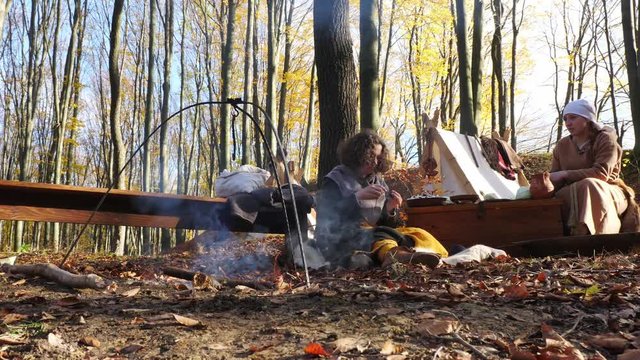 A woman hangs a cauldron on an open fire in a Viking camp.