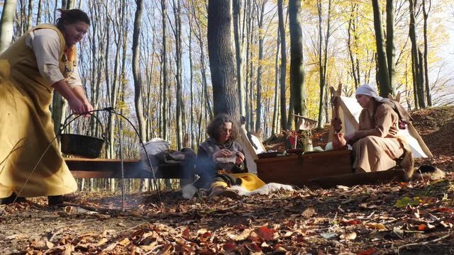 A woman hangs a cauldron on an open fire in a Viking camp.