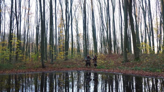 Medieval Vikings talking in the forest near lake reflected in water.