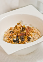 Close up, top view of a square, deep, white bowl of granola with raisins and walnut halves with skim milk on a white place mat, paleo diet