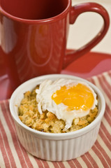 Close up, top view of a ramekin filled with freshly baked, macaroni and cheese and a poached egg sitting on a red and white stripped napkin with a red coffee cup on a round, red plate