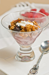 Top view, close up of a display glass of maple quinoa granola  with an artisan spoon and a dish of raspberries on a white table cloth