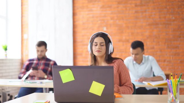 young happy brunette woman in big headphones sitting in front of laptop in office. Joyful girl dances and laughs at the desktop. In the background, young students of the mixed race learn. 4k
