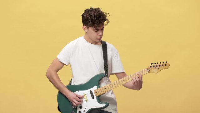 Portrait of attractive male musician with curly hair masterly playing electric guitar at show or rehearsal, isolated over yellow background
